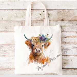 May include: A white tote bag featuring a watercolour illustration of a Highland cow wearing a floral crown. The cow is brown and white with a fluffy mane. The text "Poppets Personal Gifts" is printed on the bag.
