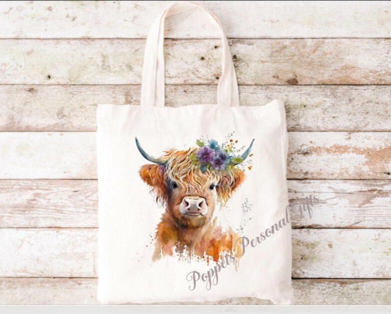 May include: A white tote bag featuring a watercolour illustration of a Highland cow wearing a floral crown. The cow is brown and white with a fluffy mane. The text "Poppets Personal Gifts" is printed on the bag.