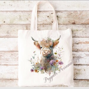 May include: A white canvas tote bag featuring a watercolour illustration of a Highland cow with flowers. The text "Poppeds Personal Gifts" is printed on the bag.