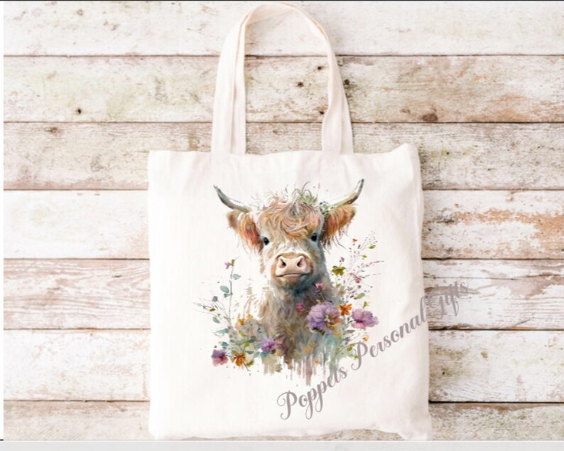 May include: A white canvas tote bag featuring a watercolour illustration of a Highland cow with flowers. The text "Poppeds Personal Gifts" is printed on the bag.