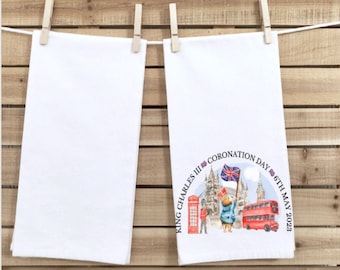 May include: White tea towel featuring a watercolour illustration for King Charles III's Coronation Day, 6th May 2023. The design includes a bear with a Union Jack, a red bus, and London landmarks.