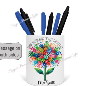 Puede incluir: Porta lápices de cerámica blanca con un diseño de huellas de manos de colores y el texto "Gracias por ser el mejor maestro de todos los tiempos". El porta lápices también tiene el nombre "Sra. Smith" impreso en la parte inferior.