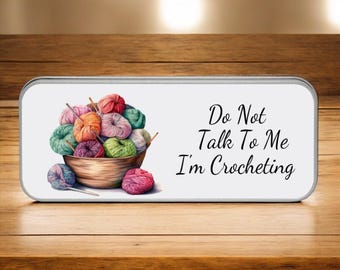 May include: A rectangular tin with a white background features a watercolor illustration of colorful yarn balls in a wooden bowl. The text "Do Not Talk To Me I'm Crocheting" is written in black script.