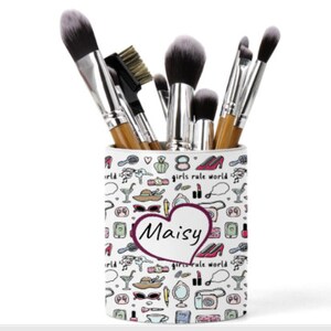 May include: White ceramic makeup brush holder with a pink heart and the name "Maisy" in a pink heart. The holder is decorated with a pattern of makeup items, shoes, and the text "girls rule world".