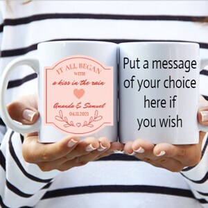 May include: White ceramic mug with a pink and red design that says "It all began with a kiss in the rain" and "Amanda & Samuel 04.11.2021". The other mug is white and says "Put a message of your choice here if you wish".