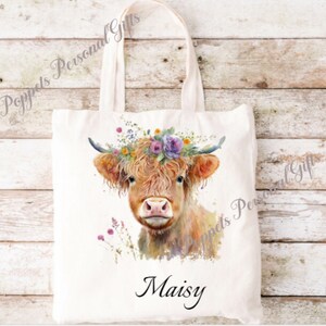 May include: A white canvas tote bag featuring a watercolour illustration of a Highland cow wearing a flower crown. The cow's name, "Maisy", is printed in black below the image.