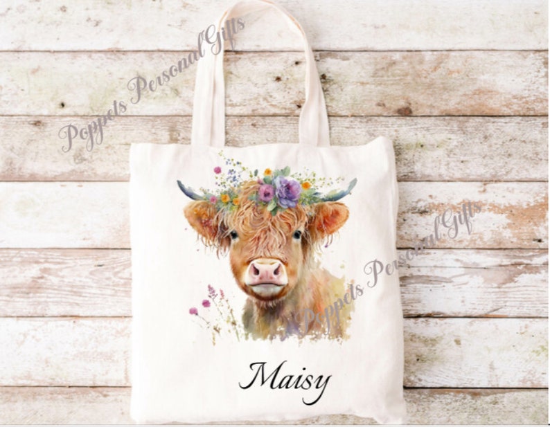 May include: A white canvas tote bag featuring a watercolour illustration of a Highland cow wearing a flower crown. The cow's name, "Maisy", is printed in black below the image.