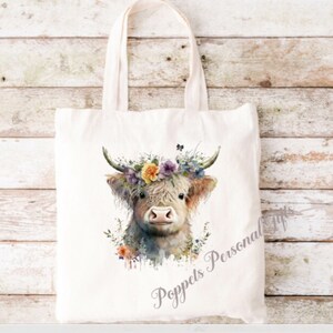 May include: A white canvas tote bag featuring a watercolour illustration of a Highland cow wearing a floral crown. The cow is looking at the viewer with a sweet expression. The text "Poppets Personal Gifts" is printed on the bag.