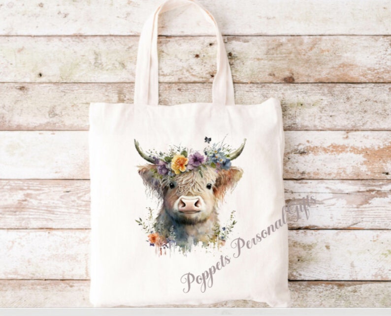 May include: A white canvas tote bag featuring a watercolour illustration of a Highland cow wearing a floral crown. The cow is looking at the viewer with a sweet expression. The text "Poppets Personal Gifts" is printed on the bag.