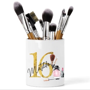 May include: White ceramic makeup brush holder with gold glitter accents. The holder features the number 16 and the name Matilda in gold glitter. The holder is filled with makeup brushes.