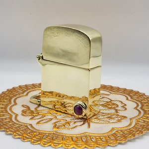 Può includere: Un accendino dorato con una pietra preziosa viola sul fondo. L'accendino è appoggiato su un pizzo bianco con un bordo dorato.