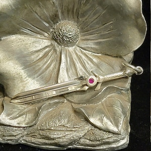 Può includere: Spilla argentata a forma di spada in miniatura con una gemma rossa sull'elsa, appoggiata su uno sfondo floreale scolpito. La spilla presenta dettagli intricati e una finitura lucida, suggerendo un accessorio decorativo.