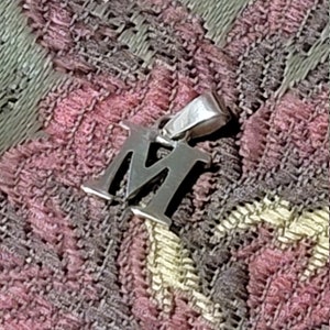 May include: A silver charm in the shape of the letter M. The charm is on a pink and gray patterned fabric.