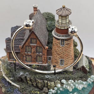 May include: A silver-toned bangle bracelet with spherical end caps is displayed in front of a detailed miniature lighthouse scene. The scene features a brick lighthouse, a house, and a rocky coastline with ocean waves.