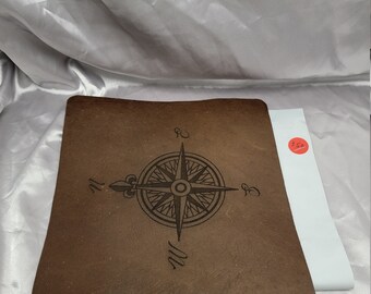 May include: Brown leather desk mat with a compass design engraved in the centre. The compass has a fleur-de-lis at the top and the cardinal directions are labelled N, E, S, and W.