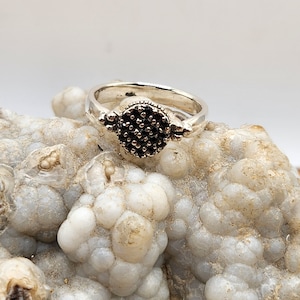 May include: A silver ring featuring a round black stone set in the centre. The ring has a textured band and is accented with small silver beads.
