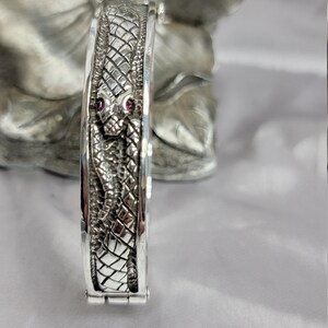 May include: A silver snake bangle bracelet with two ruby red stones for eyes. The bracelet is detailed with a snake skin pattern.
