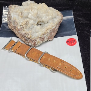 May include: A brown leather watch band with silver metal clasps. The band is on a white surface with a clear plastic bag and a white rock with brown markings.