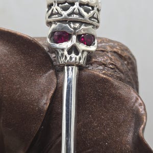 May include: A silver skull with a crown on top and red gemstone eyes. The skull is attached to a silver rod.