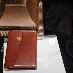May include: A small, brown leather wallet with white stitching and a brass button closure. The wallet is rectangular and appears to be handmade. The background includes a decorative lamp and a white tag with the number 38.
