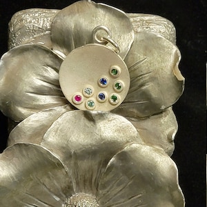 May include: A silver pendant with a round, textured surface and a cluster of colorful gemstones, including red, blue, green, and white.