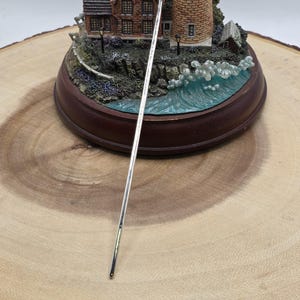 May include: A silver metal bottle stopper with a round top and a long, slender stem. The stopper is displayed on a wooden surface, with a miniature lighthouse and house diorama in the background.