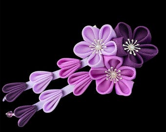 Puede incluir: Accesorio para el cabello floral artesanal con tres flores en tonos morados y lavanda, con delicados detalles de pétalos y una cuenta central. El accesorio también incluye acentos de cinta en cascada en colores coordinados, sobre un fondo negro.