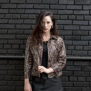 K&ouml;nnte beinhalten: Eine Frau tr&auml;gt eine Jacke mit Schlangenhautmuster, ein schwarzes Oberteil, einen Jeans-Minirock und Schlangenhaut-Schaftstiefel. Sie steht vor einer schwarzen Backsteinwand.