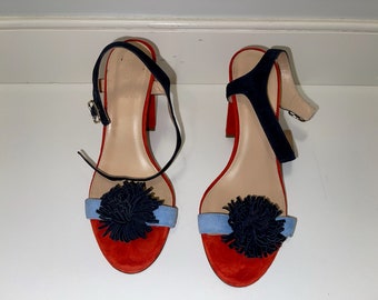 May include: A pair of red suede heeled sandals with blue suede straps and navy blue pom-poms.