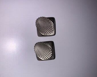 May include: A pair of silver tone earrings with a seashell design. The earrings are square shaped with a raised seashell design in the center.