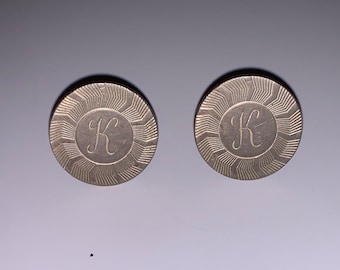 May include: Two silver cufflinks with a sunburst design and the letter "K" engraved in the center.