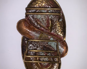 May include: A large, oval-shaped belt buckle made of brass with a textured, hammered finish. The buckle has a reddish-brown patina and features a raised, abstract design. The buckle has a rectangular, brass-plated metal strap.