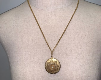 May include: A gold chain necklace with a round locket pendant. The locket is decorated with a gold filigree design and a single pearl in the center.