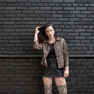 K&ouml;nnte beinhalten: Eine Frau tr&auml;gt eine braune und schwarze Jacke mit Schlangenmuster, ein schwarzes Oberteil, einen schwarzen Minirock und schwarze und braune gemusterte Strumpfhosen. Sie steht vor einer schwarzen Backsteinwand.