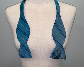 May include: A teal blue bow tie with pink and navy blue stripes. The bow tie is on a white mannequin.