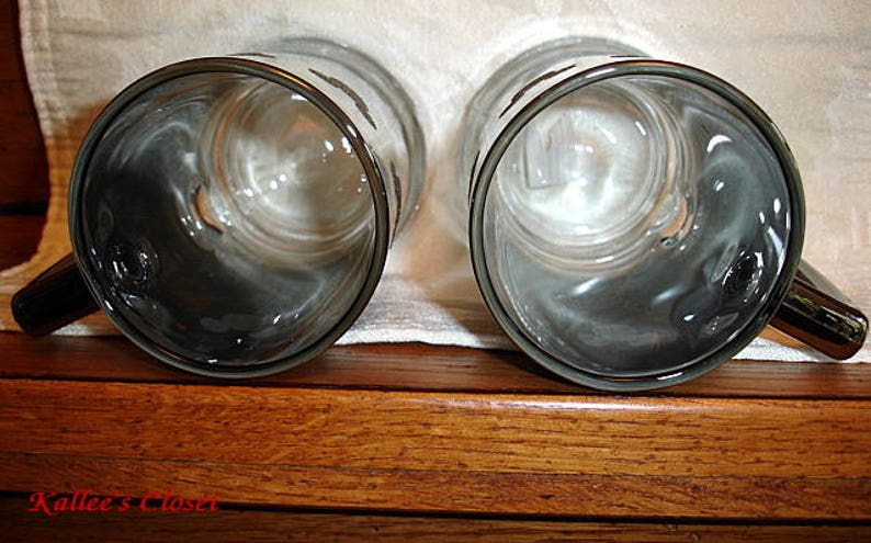 May include: Two clear glass mugs with black rims and handles. The mugs are empty and sitting on a wooden surface.