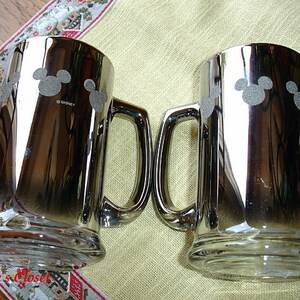 May include: Two silver metallic beer mugs with a Mickey Mouse silhouette design. The mugs have a handle and are made of glass.