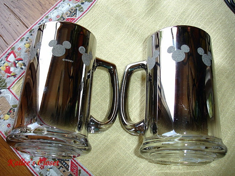 May include: Two silver metallic beer mugs with a Mickey Mouse silhouette design. The mugs have a handle and are made of glass.