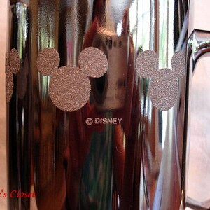 May include: A silver metal mug with a black handle. The mug has a design of three Mickey Mouse heads with a brown glitter finish. The mug has the text "&copy;DISNEY" printed on the bottom.