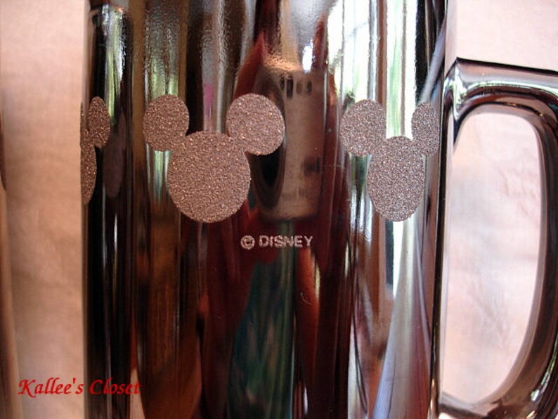 May include: A silver metal mug with a black handle. The mug has a design of three Mickey Mouse heads with a brown glitter finish. The mug has the text "&copy;DISNEY" printed on the bottom.