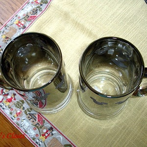 May include: Two silver-rimmed glass mugs with a decorative design. The mugs have a clear glass body and a silver rim around the top.