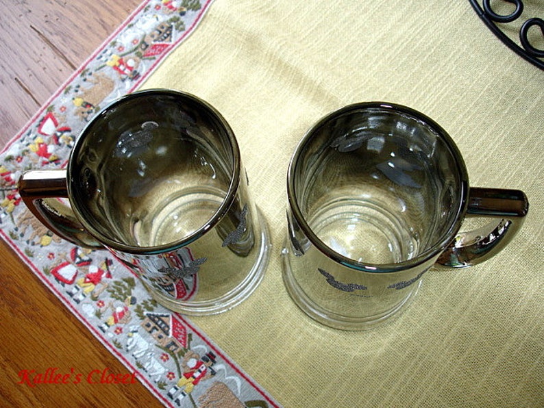 May include: Two silver-rimmed glass mugs with a decorative design. The mugs have a clear glass body and a silver rim around the top.