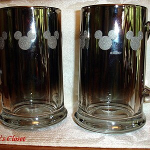 May include: Two silver metallic beer mugs with a Mickey Mouse silhouette design. The mugs have a handle and are etched with the word "Disney".