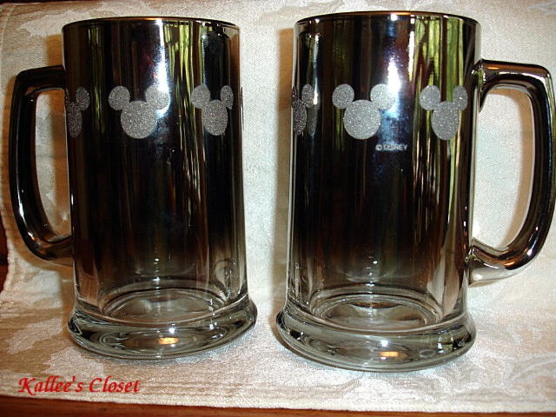 May include: Two silver metallic beer mugs with a Mickey Mouse silhouette design. The mugs have a handle and are etched with the word "Disney".