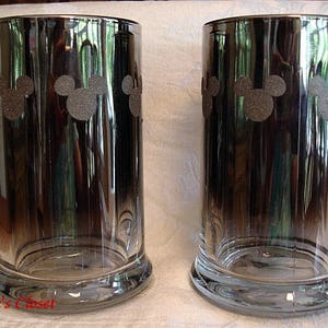 May include: Two clear glass mugs with a black and silver ombre effect. Each mug has a silhouette of Mickey Mouse's head repeated around the mug.