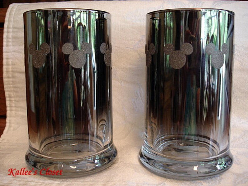 May include: Two clear glass mugs with a black and silver ombre effect. Each mug has a silhouette of Mickey Mouse's head repeated around the mug.