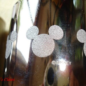 May include: Silver metal tumbler with a Mickey Mouse design. The tumbler has a shiny, reflective surface and the Mickey Mouse design is in a silver color.