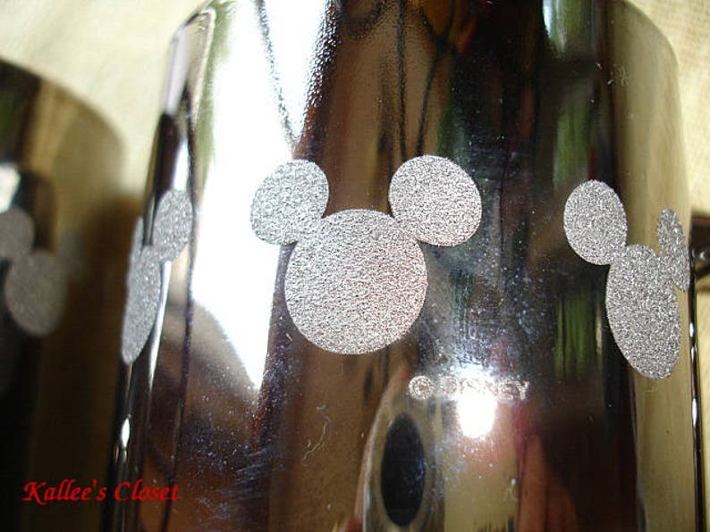 May include: Silver metal tumbler with a Mickey Mouse design. The tumbler has a shiny, reflective surface and the Mickey Mouse design is in a silver color.