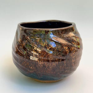May include: A rounded ceramic bowl featuring a glossy, dark brown glaze with swirling metallic bronze and deep blue undertones. The exterior surface has a textured, undulating finish with visible indentations, while the interior is smooth and dark.