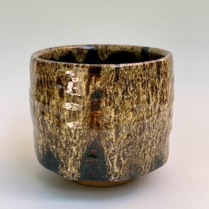 May include: A cylindrical ceramic cup featuring a reactive glaze with a dark brown base and a textured, drippy cream-colored overlay. The exterior surface displays vertical streaks and mottled patterns, while the interior is a solid dark brown.